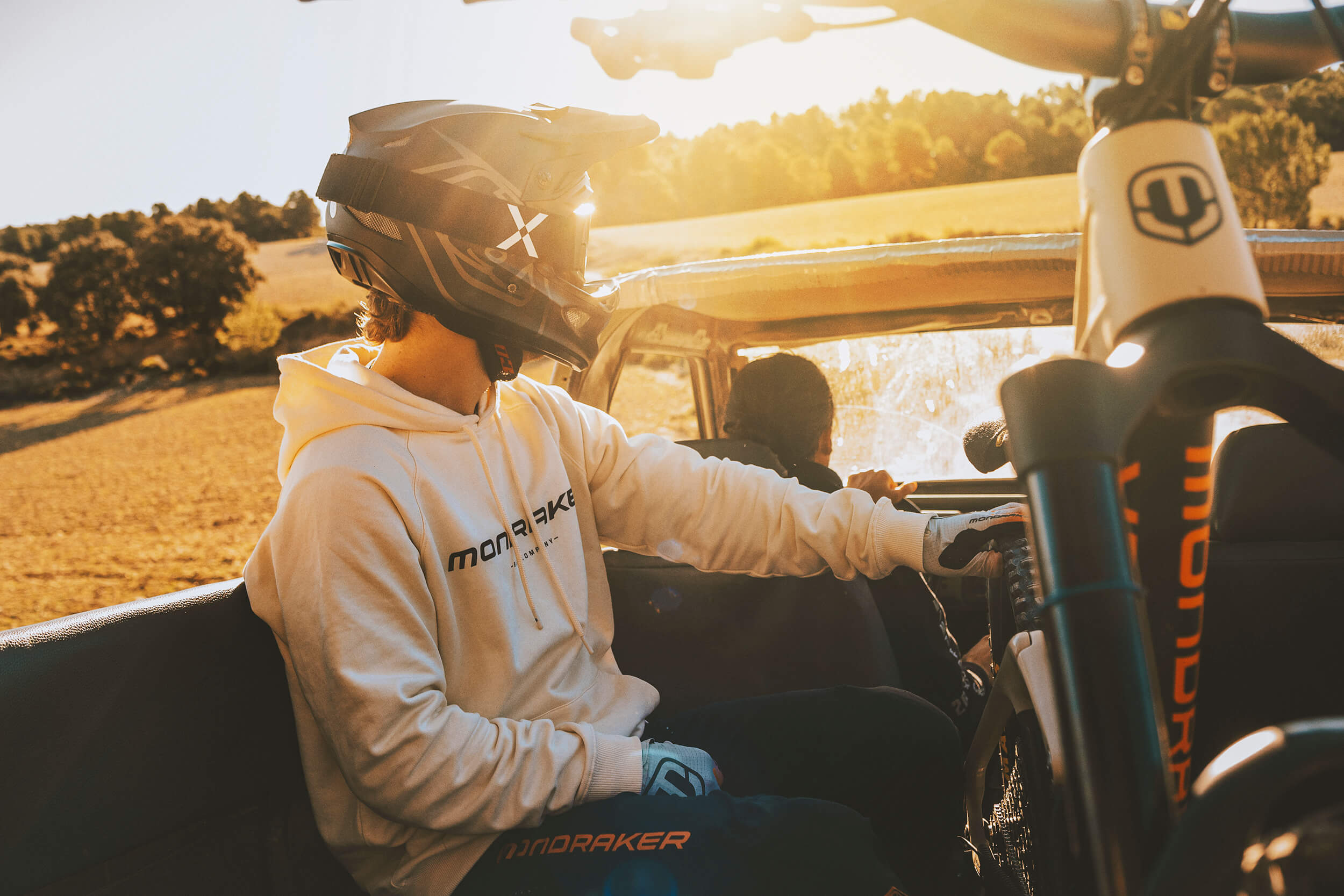 Person in Mondraker Hoodie und Helm, im Auto mit Mountainbike, sonnige Landschaft im Hintergrund. Geeignet für Outdoor-Sport.