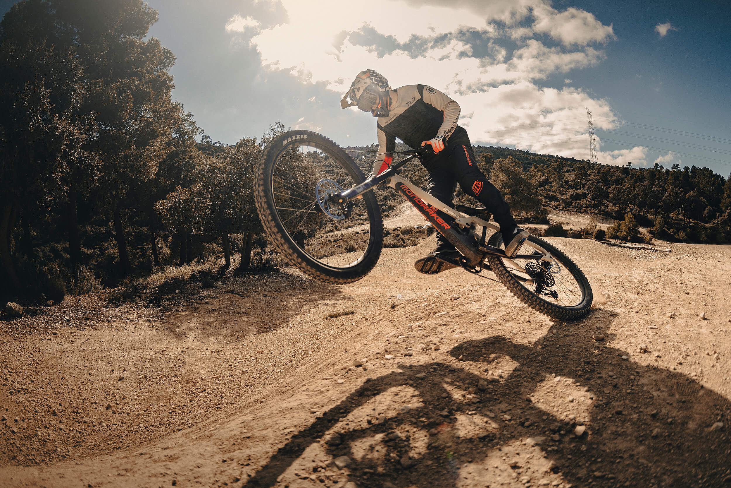 Person auf Mondraker-Mountainbike im Gelände bei Sprung, sonniger Tag, geeignet für Mountainbike-Enthusiasten.