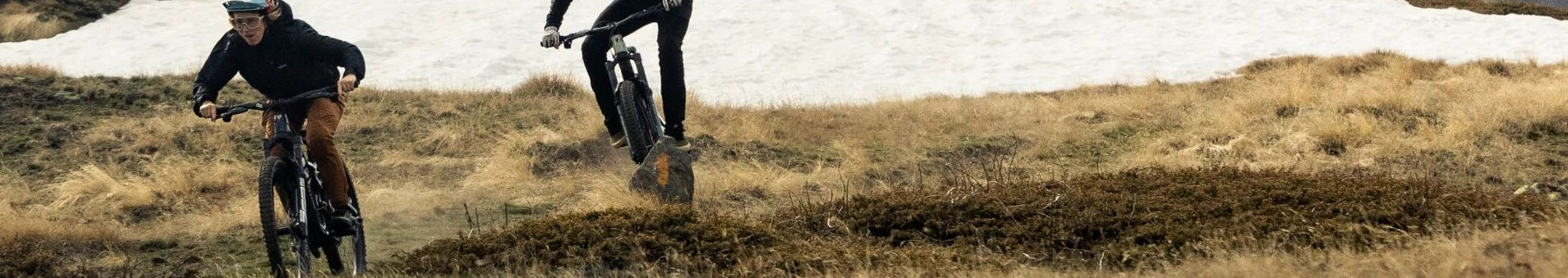 Zwei Personen fahren Trek E-MTBs auf einem grasigen Pfad mit schneebedecktem Hintergrund.