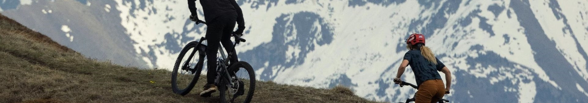Zwei Personen fahren mit Mountainbikes einen Bergpfad hinauf, im Hintergrund schneebedeckte Berge.
