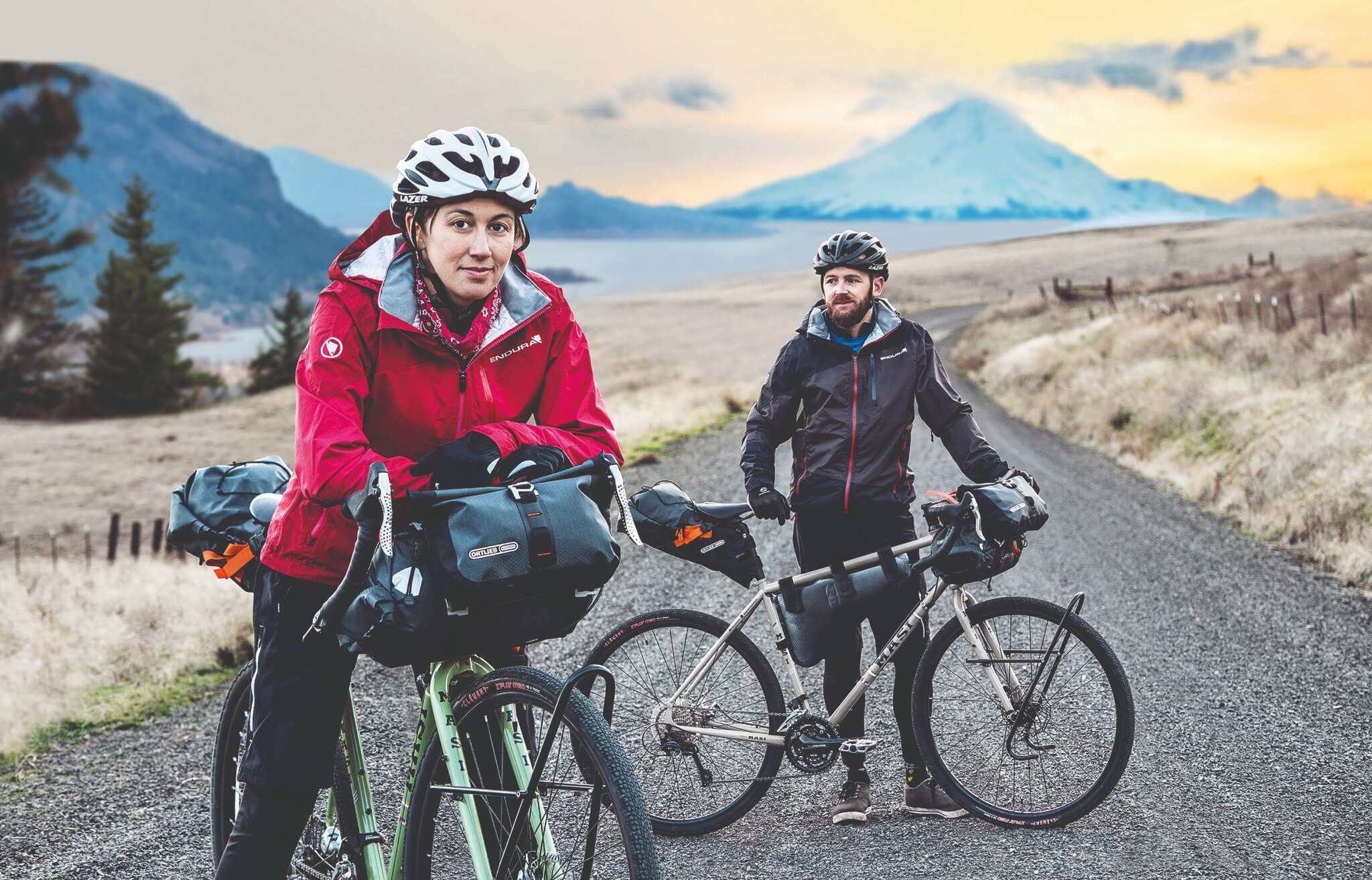 Zwei Radfahrer auf Gravelbikes mit Ortlieb-Taschen, ausgerüstet für Abenteuerreisen, auf einer Kiesstraße vor Bergkulisse.