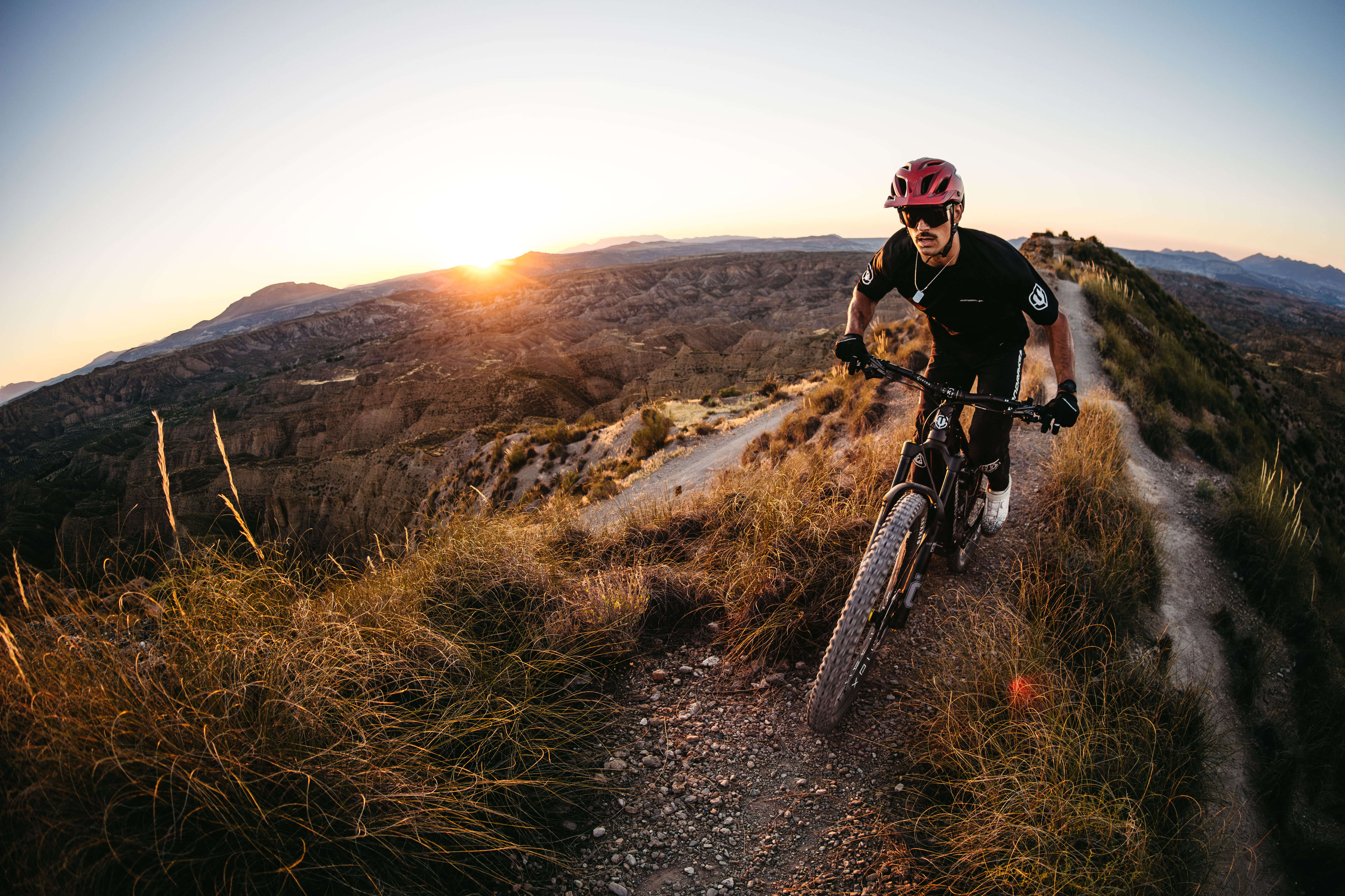 Mountainbiker auf einem Mondraker E-Bike fährt bei Sonnenuntergang auf einem rauen Pfad in malerischer Berglandschaft.