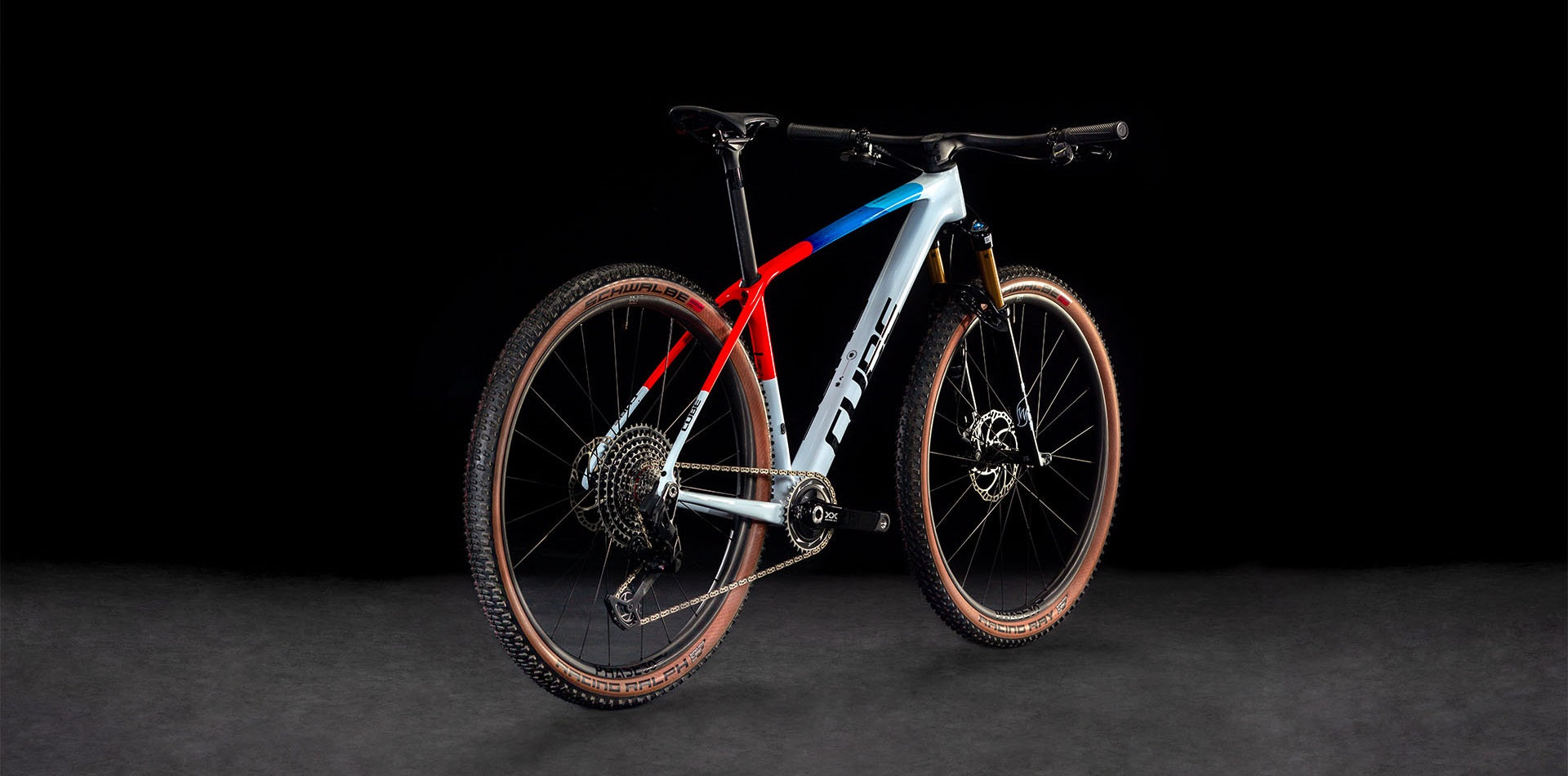 Cube Mountainbike mit rotem, blauem und grauem Rahmen, Schwalbe-Reifen und goldener Federgabel, auf schwarzem Hintergrund.