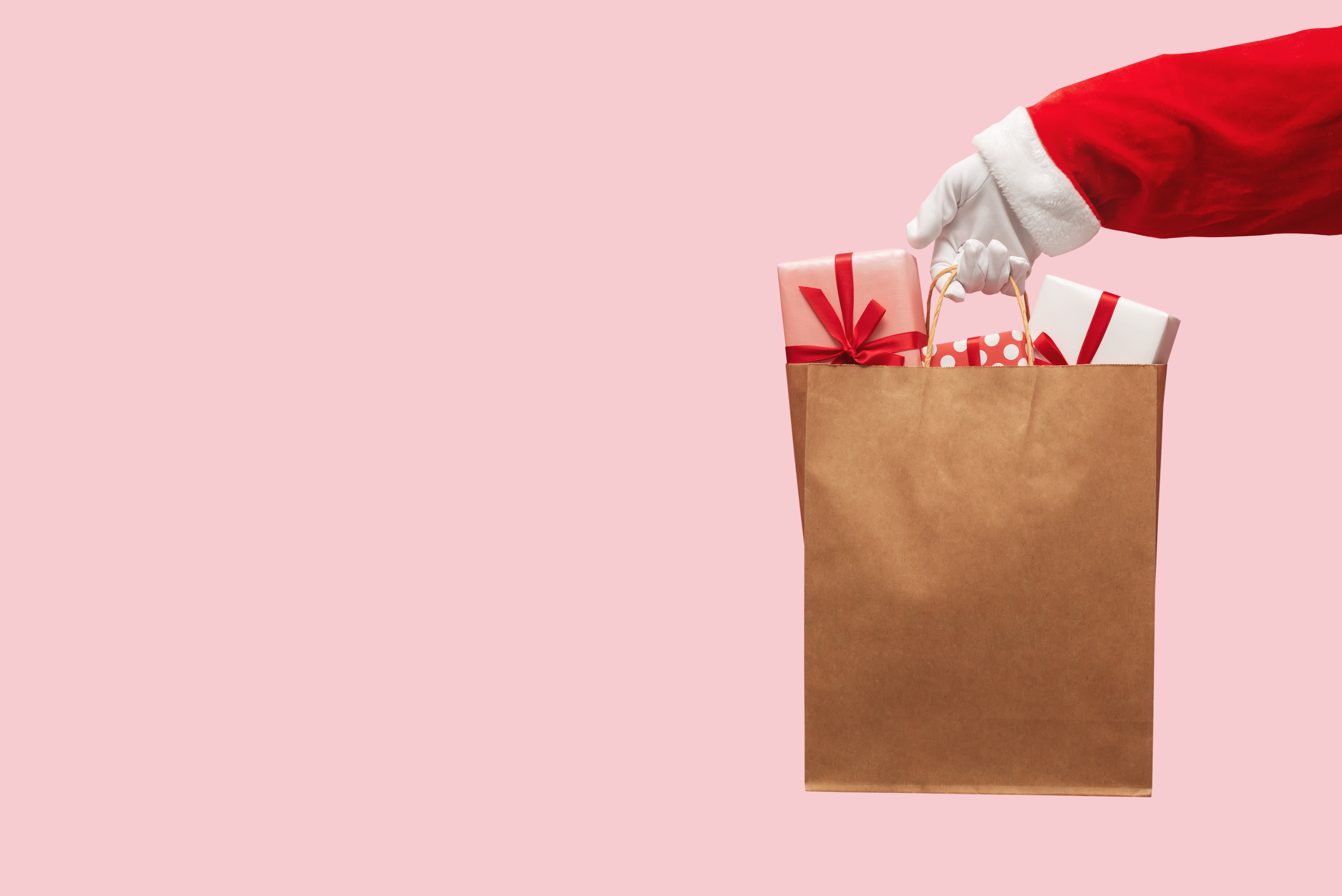 Weihnachtliche Tüte mit Geschenken, von Hand im roten Ärmel gehalten, vor rosa Hintergrund.