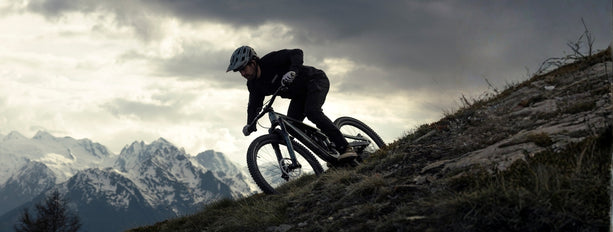 Radfahrer fährt Downhill auf einem Mountainbike im Gebirge, schneebedeckte Berge im Hintergrund.