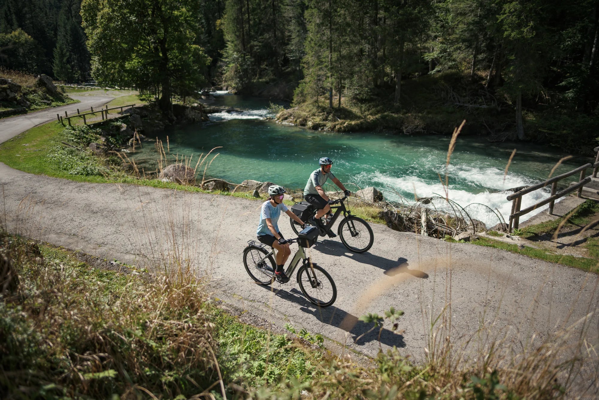 Zwei Personen radeln auf E-Bikes entlang eines Flusses in einer idyllischen Waldlandschaft.