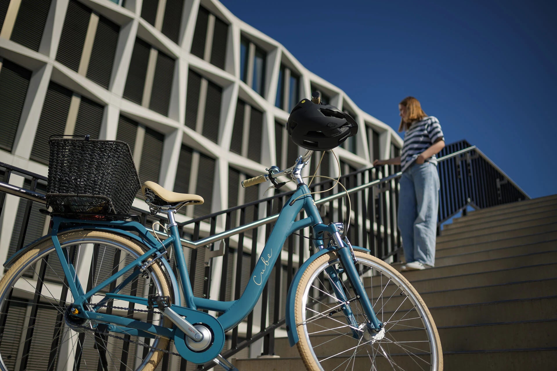 Blaues Cube-Damenfahrrad mit Korb, abgestützt an einer Treppe. Junge Frau mit gestreiftem Oberteil im Hintergrund.