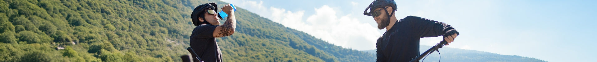 Zwei Personen auf Mountainbikes mit Helmen; eine trinkt, die andere hält den Lenker; bewaldete Hügel im Hintergrund.