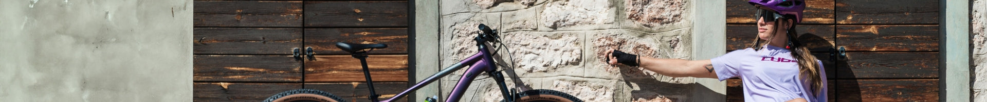 Frau in lila Cube-Shirt mit Fahrrad und Helm vor Steinwand; ideal für sportliche Frauen und Outdoor-Aktivitäten.
