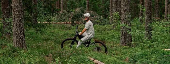 Person auf einem Mountainbike fährt durch einen Wald; trägt Helm und Freizeitsportkleidung, umgeben von Bäumen und Pflanzen.