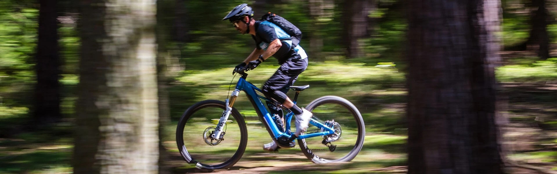 Person fährt auf blauem E-Mountainbike durch Wald. Helm und Rucksack für All-Mountain-Trail-Abenteuer.