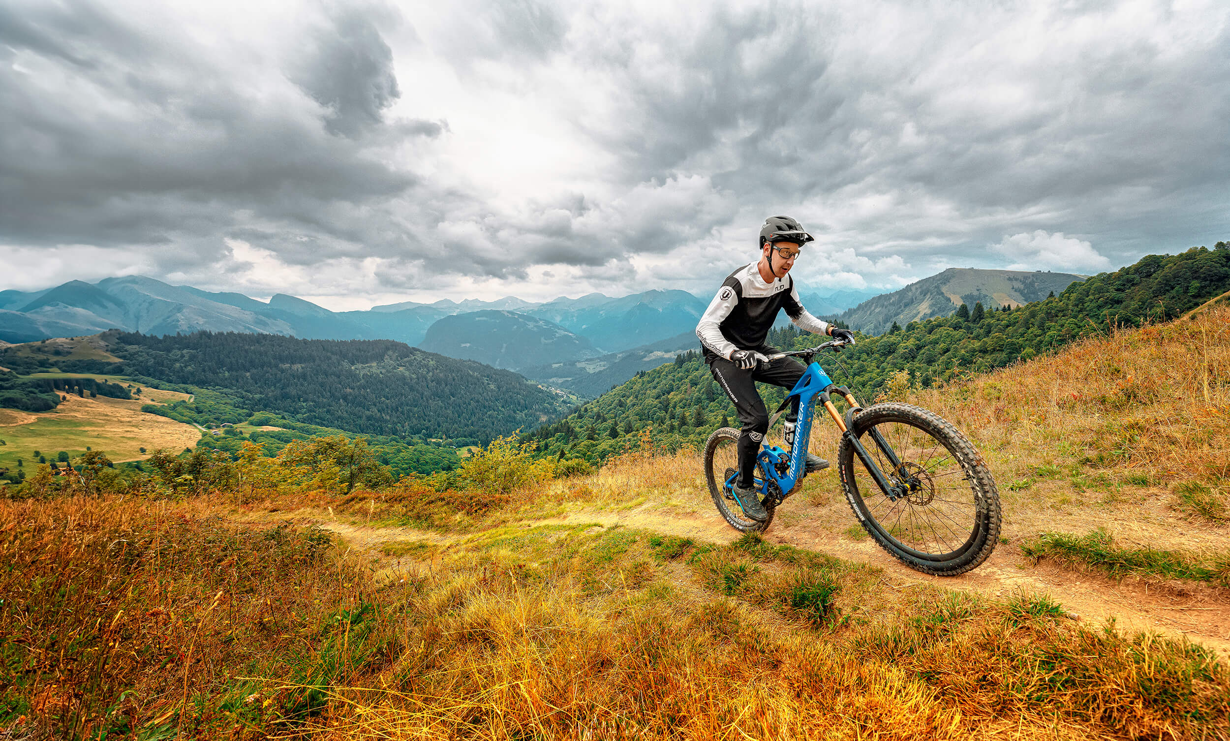 Mountainbiker fährt Mondraker Fahrrad auf einem Trail mit Bergblick im Hintergrund.