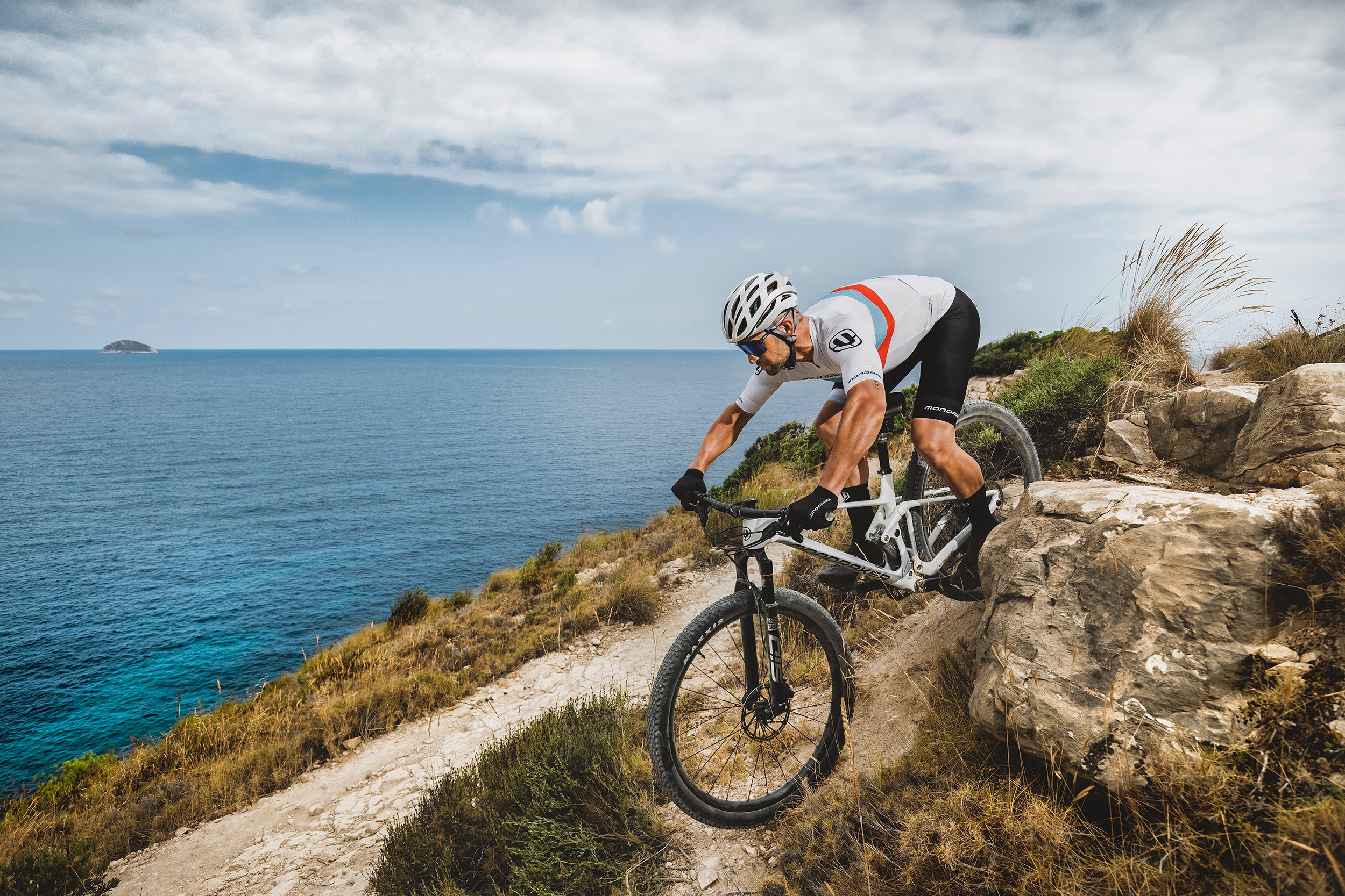 Mountainbiker auf Mondraker F-Podium fährt auf felsigem Küstenpfad. Ozeanhintergrund mit klarem, blauem Himmel.