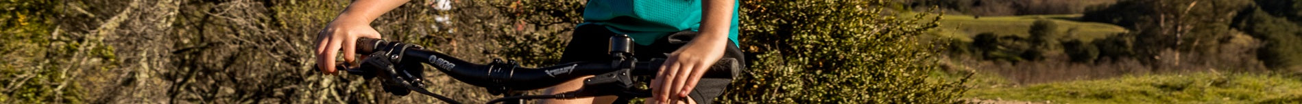 Junge Person fährt Fahrrad im Freien, hält den Lenker mit beiden Händen, grüne Vegetation im Hintergrund.