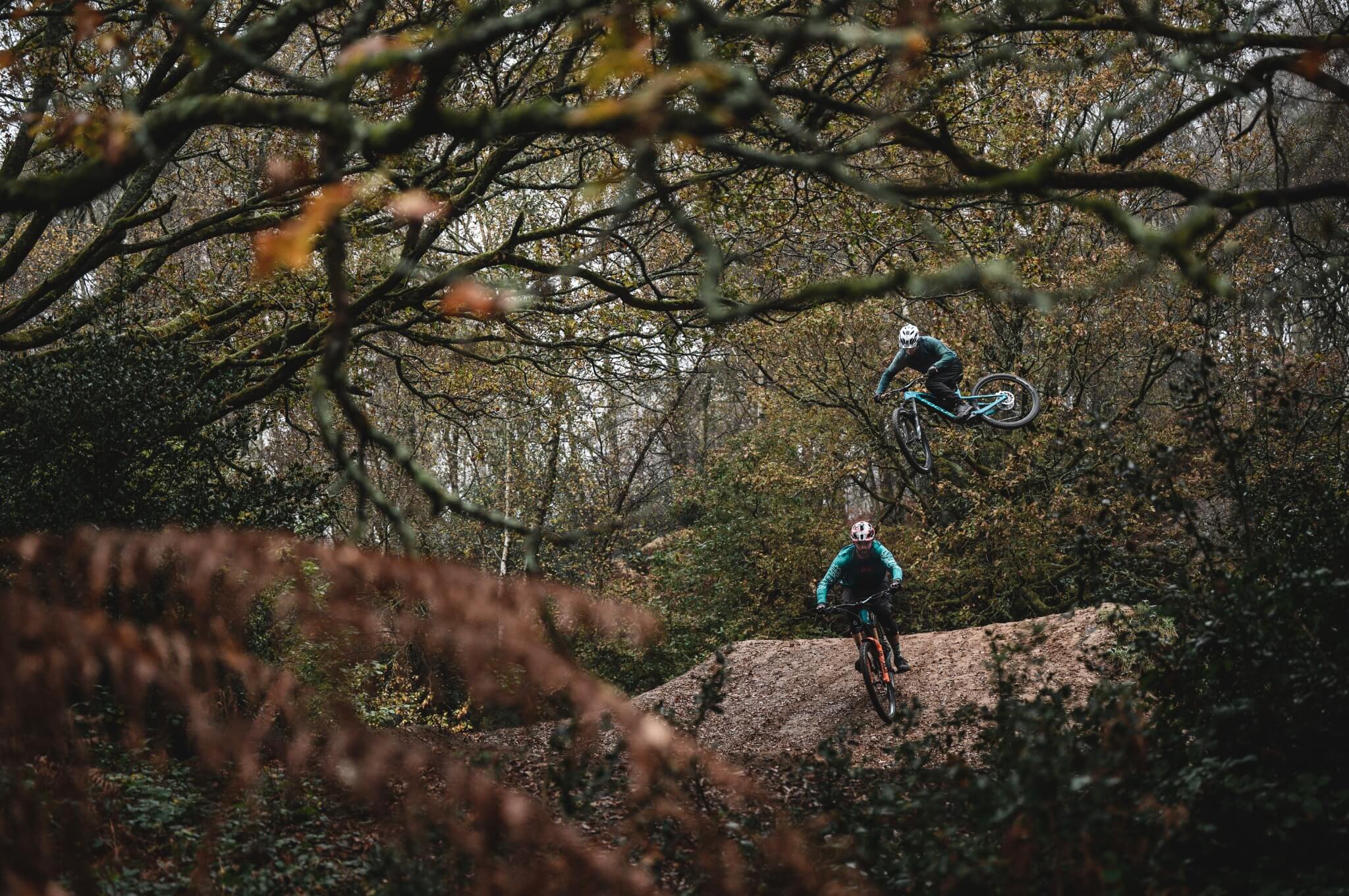 Zwei Mountainbiker in blauer Kleidung fahren durch bewaldetes Gebiet; einer springt in der Luft, der andere am Boden.