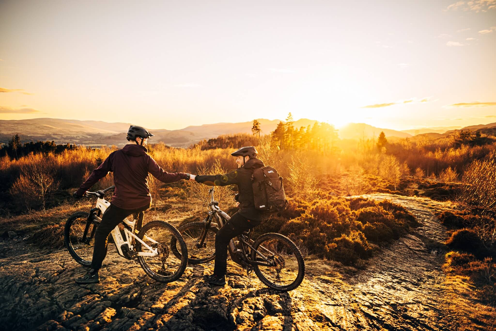 Zwei Radfahrer auf Mountainbikes im Sonnenuntergang auf einem Naturpfad, Helme und Outdoor-Kleidung tragend.