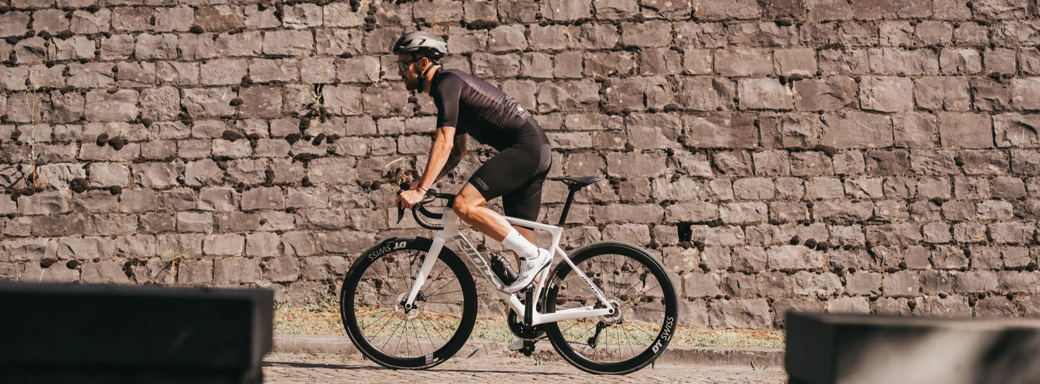 Mann fährt auf einem Ridley-Rennrad vor einer Steinmauer, in sportlicher Radkleidung und Helm.