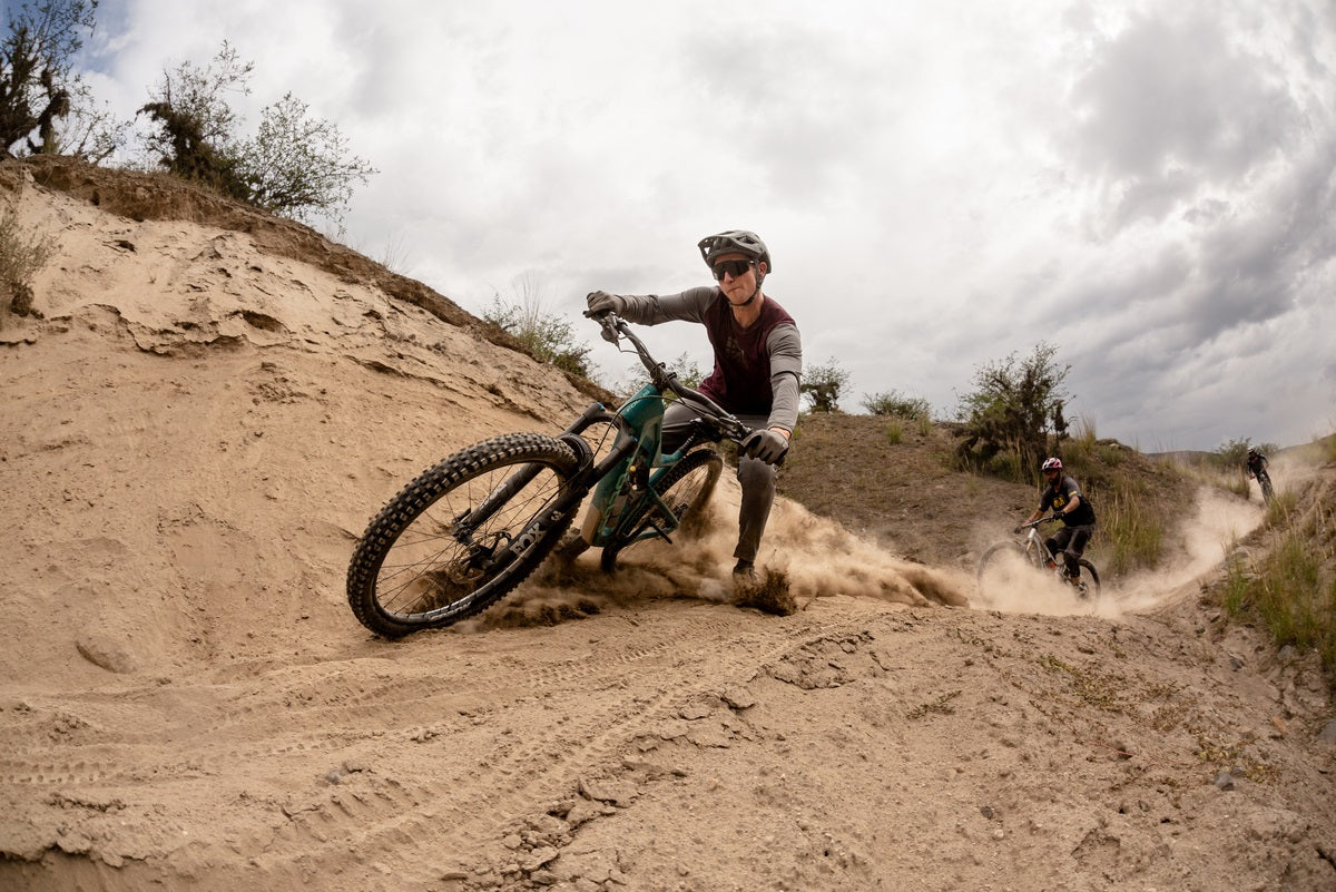 Mountainbiker fährt auf Focus Jam Fully durch Sandkurve, gefolgt von zwei weiteren Fahrern. Abenteuer und Outdoor-Sport.