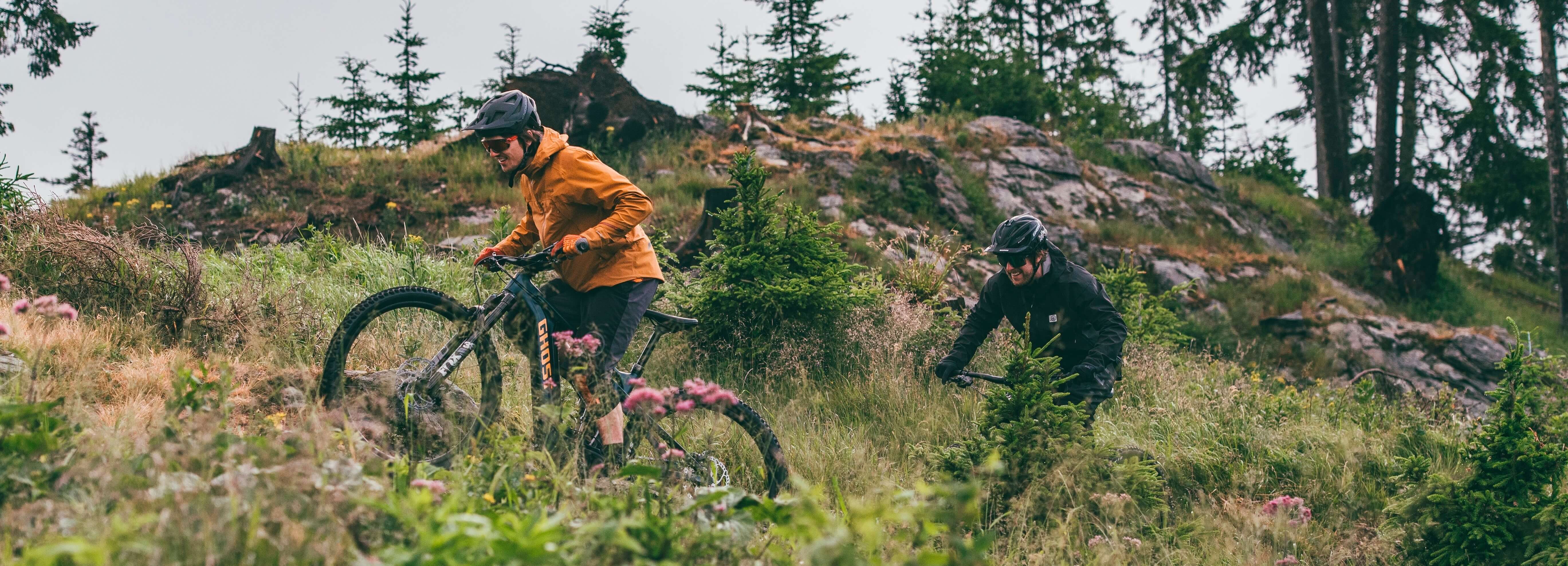 Zwei Mountainbiker fahren mit Ghost-Bikes einen steinigen Hang hinunter, umgeben von grüner Natur und Bäumen.