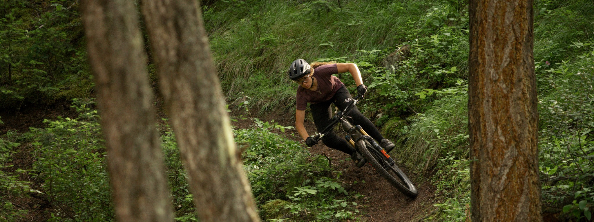 Person fährt Mountainbike auf Waldpfad zwischen Bäumen, trägt Helm und Brille für Offroad-Abenteuer.
