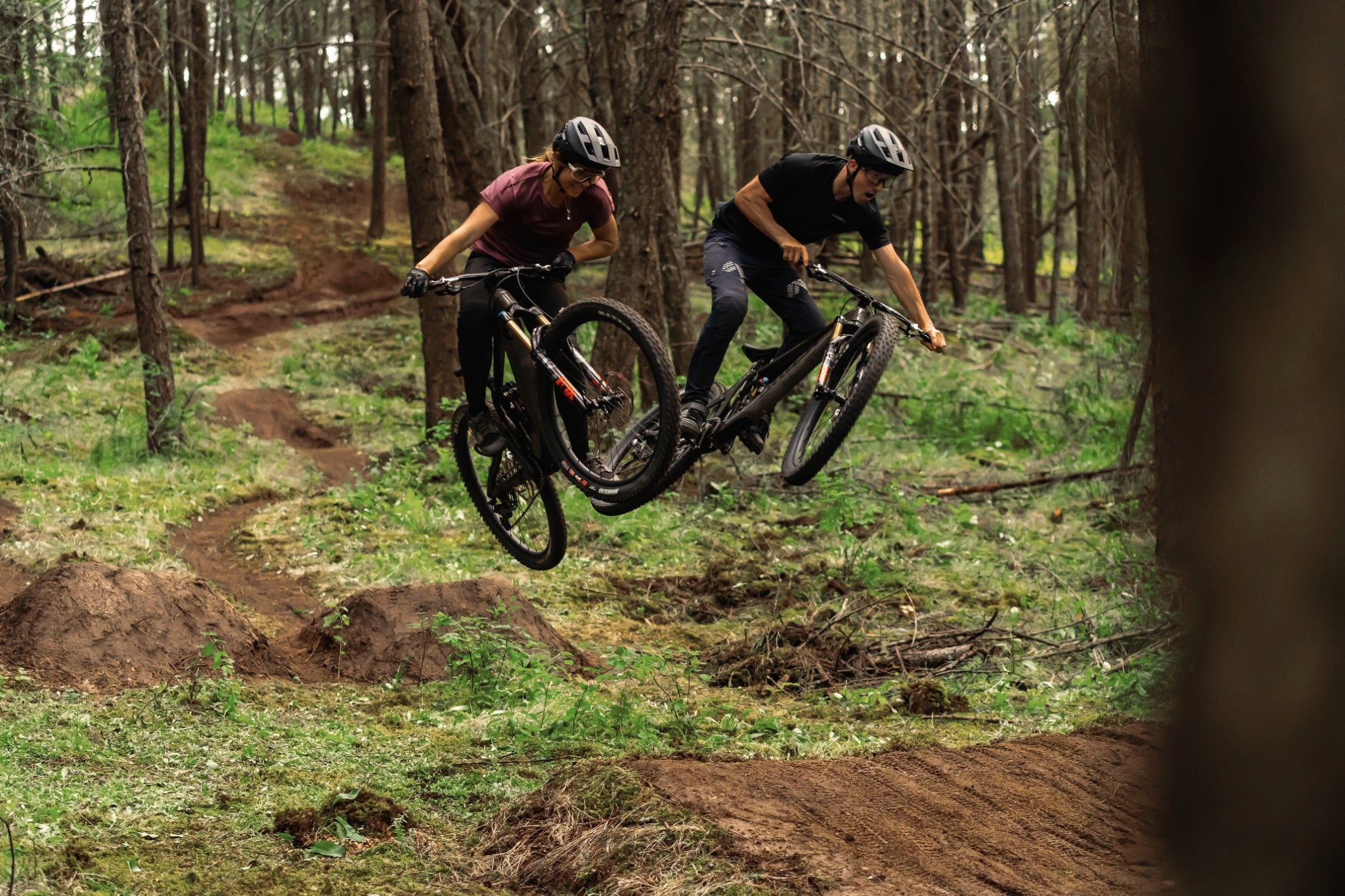 Zwei Mountainbiker springen auf einem Waldpfad. Sie tragen Helme und nutzen vollgefederte Bikes für Offroad-Fahrten.