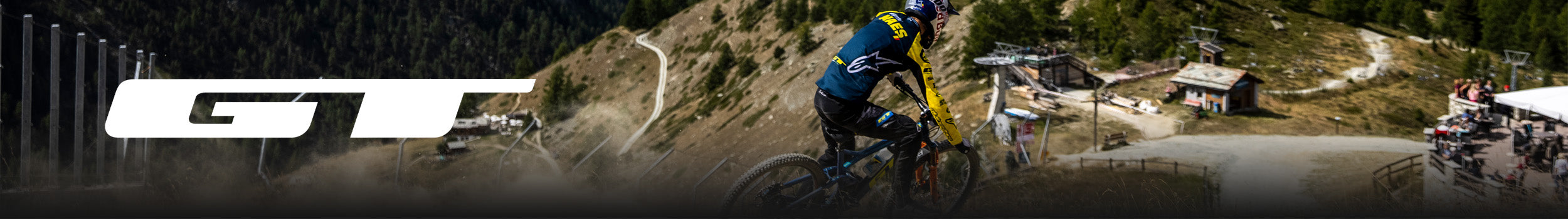 Mountainbiker fährt steilen Abhang in bergiger Landschaft hinunter, GT-Logo prominent im Bildhintergrund sichtbar.