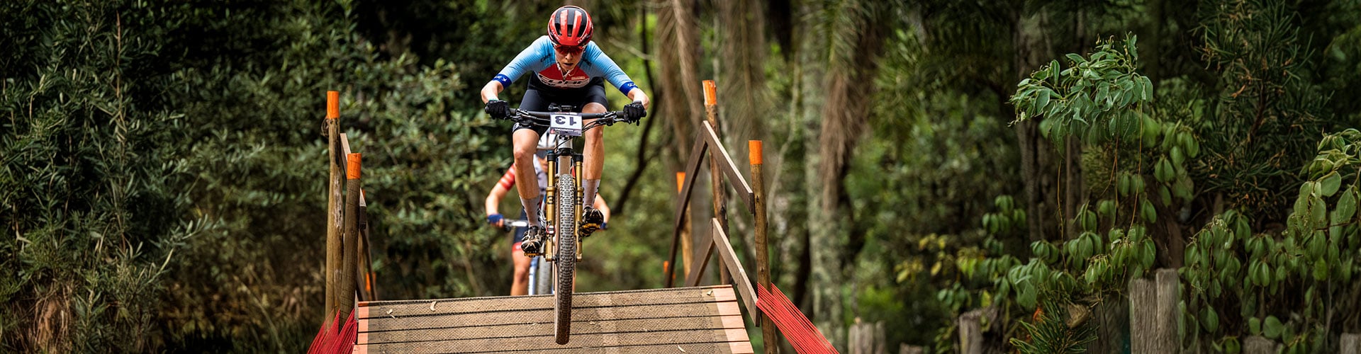 Zwei Mountainbiker bei einem Rennen über eine hölzerne Brücke im Wald, Fahrer mit rotem Helm und blauer Ausrüstung vorne.