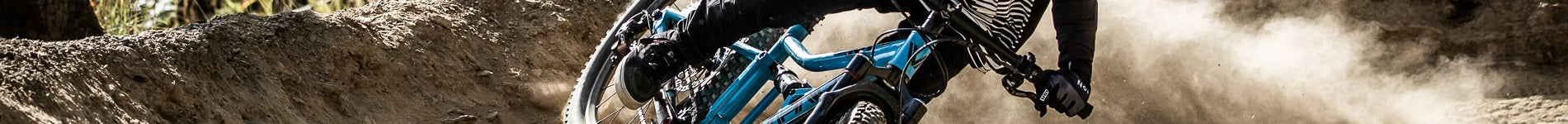 Radsportler auf einem blauen Mountainbike fährt durch staubigen Trail im Sonnenlicht, nur Räder und Beine sichtbar.
