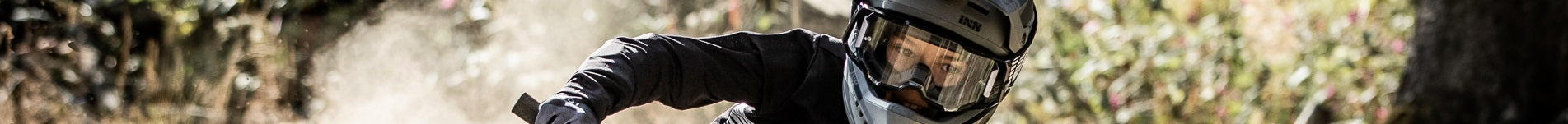 Person mit Helm und Schutzbrille auf einem Mountainbike, fährt durch Wald. Dynamische Action-Szene.