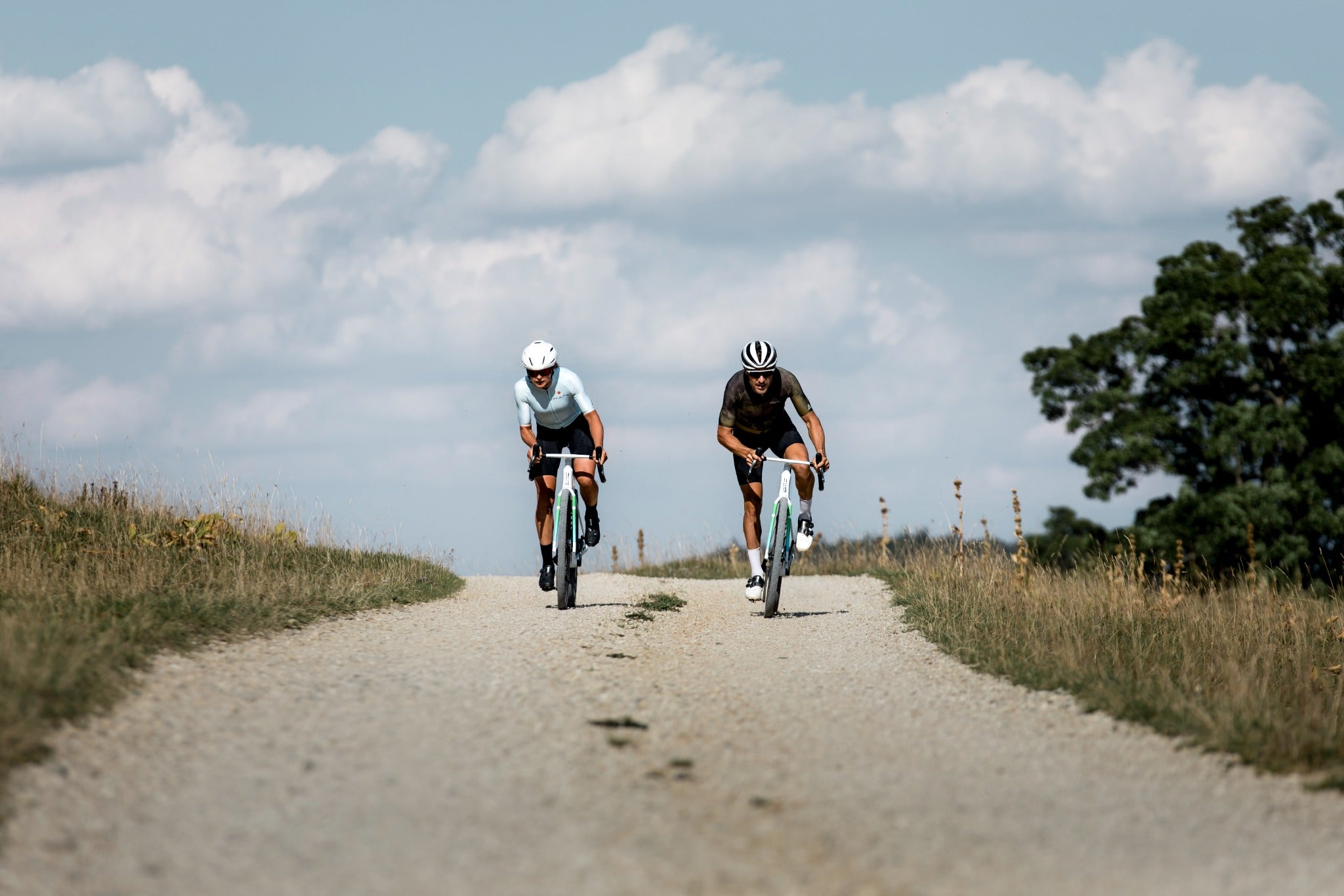 Zwei Radfahrer auf Schotterstraße bei sonnigem Wetter, mit sportlicher Kleidung und Helmen unterwegs.