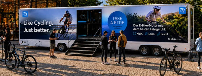 Großer weißer Lkw mit TQ-Werbung für E-Bike-Motoren, umgeben von Personen und Fahrrädern auf einer Pflasterfläche.