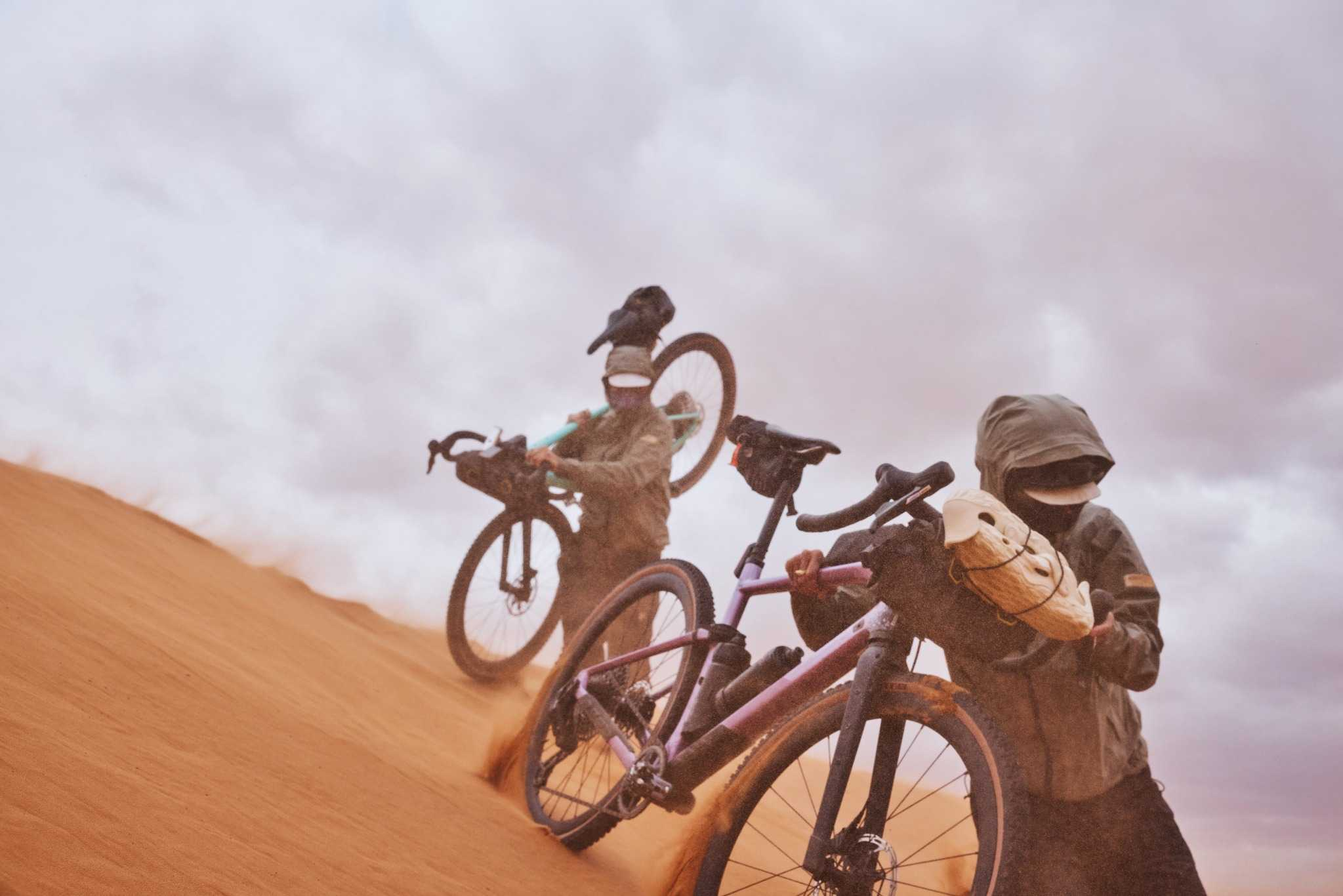 Zwei Personen tragen Fahrräder durch eine sandige Wüstenlandschaft, Modell BMC URS geeignet für Offroad-Abenteuer.
