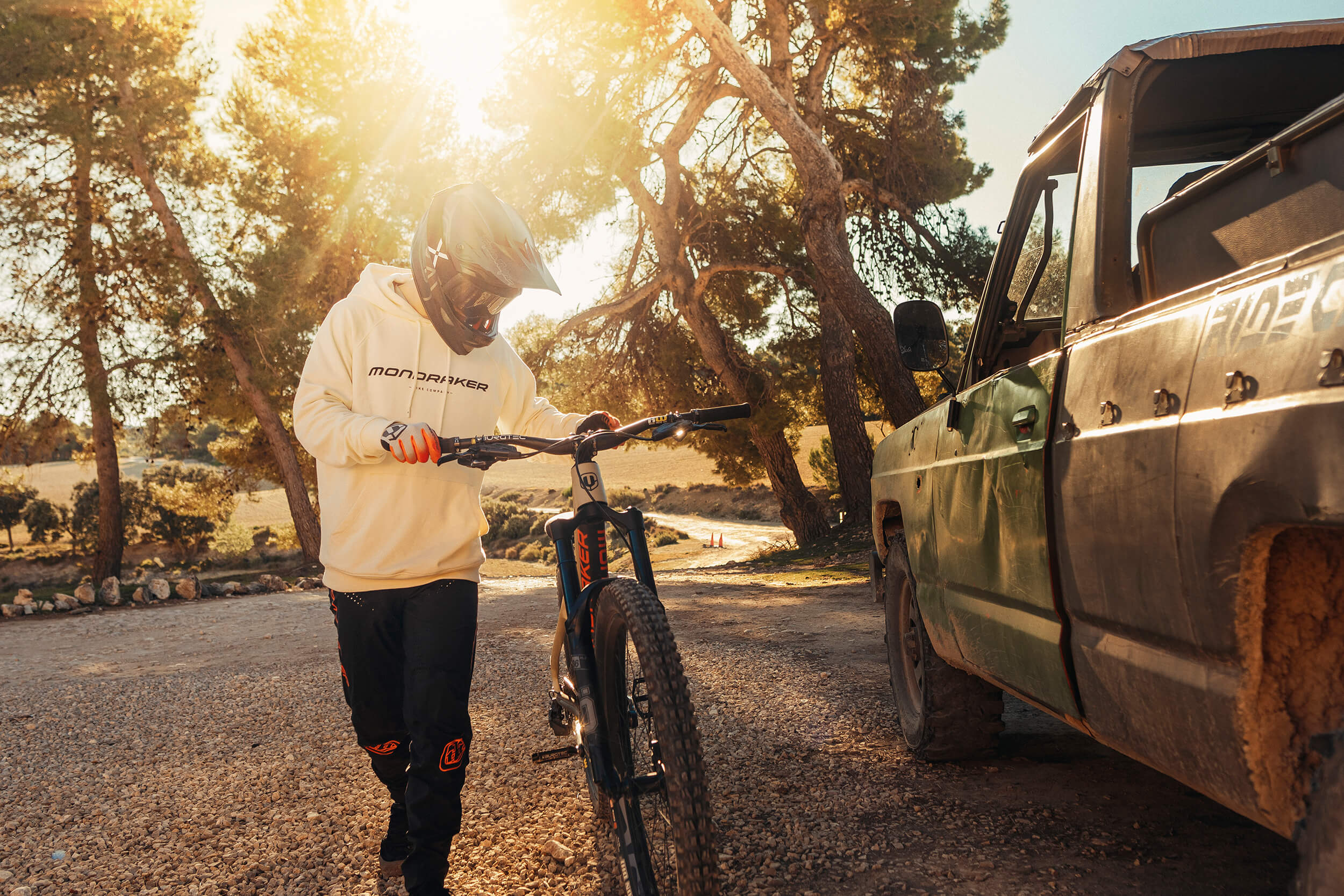 Person in Mondraker-Hoodie mit Helm hält Mountainbike, steht neben einem Geländewagen auf einem Schotterweg bei Sonnenuntergang.