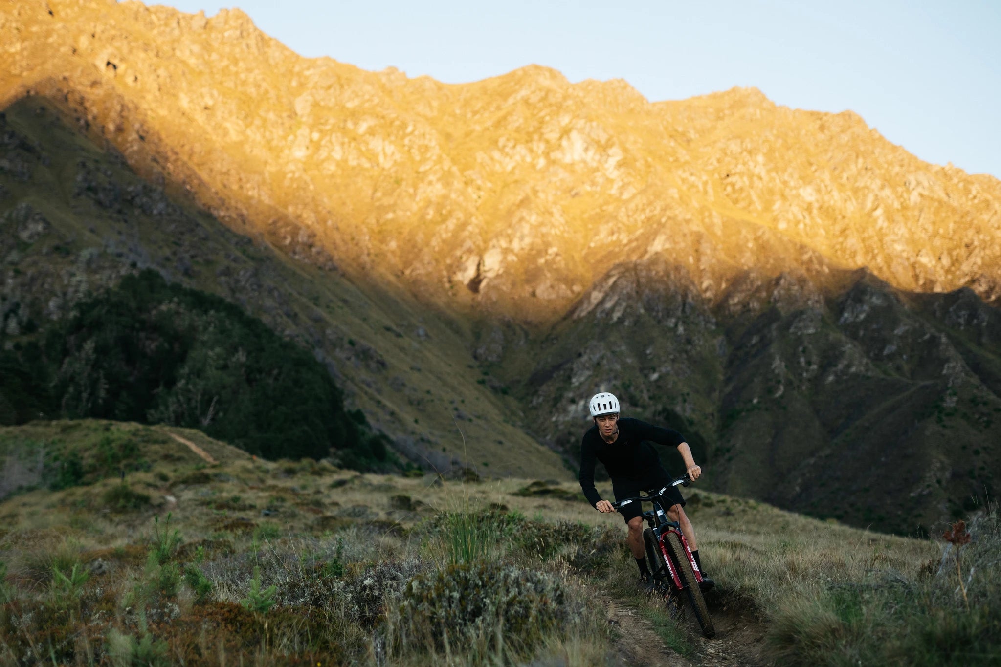Mountainbiker mit weißem Helm fährt auf einem schmalen Pfad in bergiger Landschaft bei Sonnenuntergang.