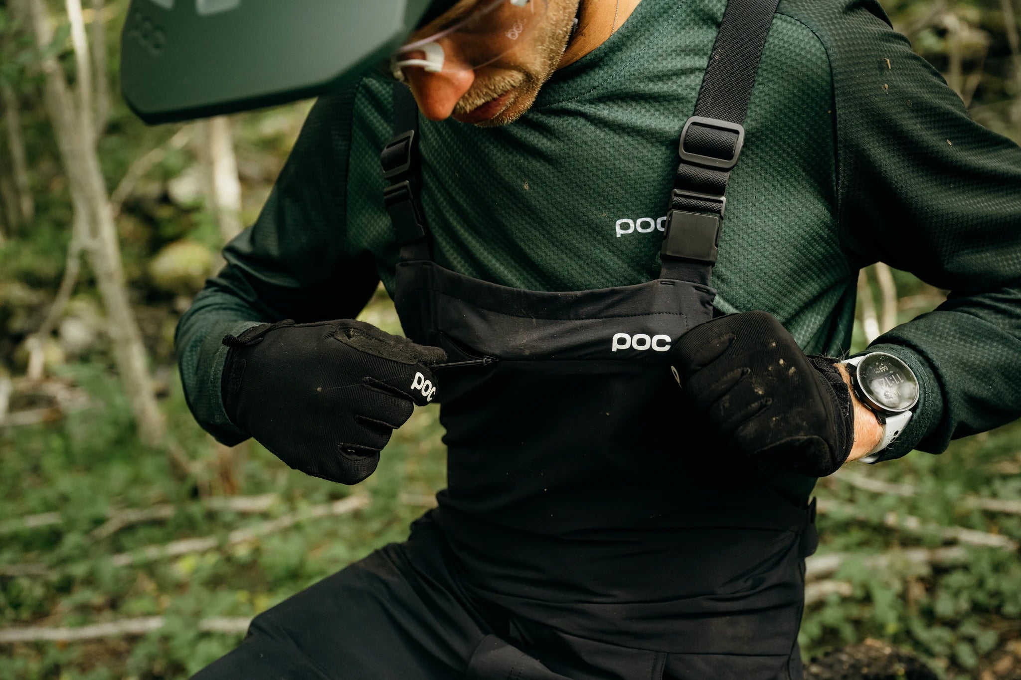 Mann in POC-Mountainbike-Bekleidung mit Handschuhen im Wald, öffnet den Reißverschluss seiner schwarzen Latzhose.