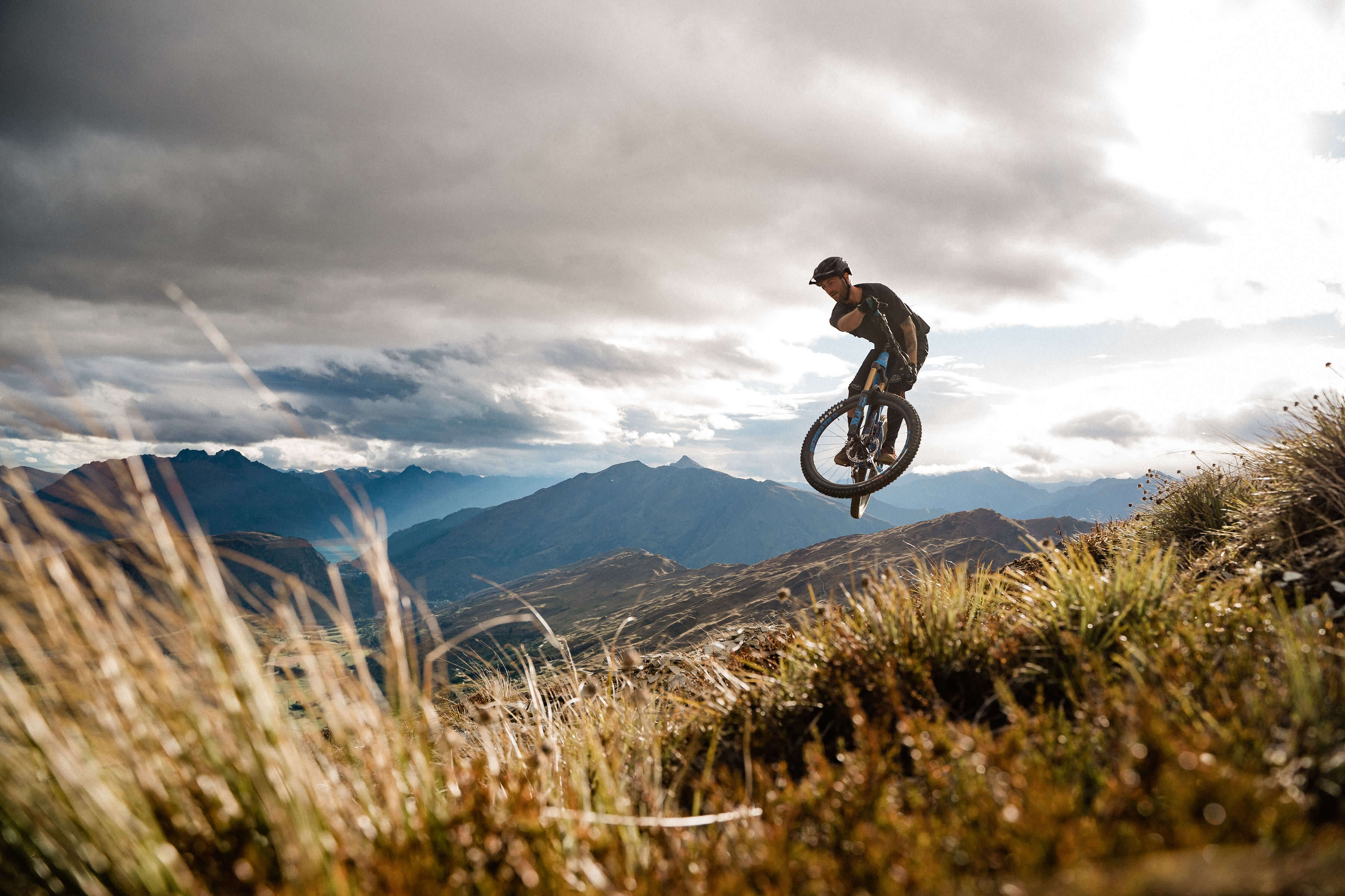 Mountainbiker springt über Hügel in bergiger Landschaft mit dramatischem Himmel, ideal für Outdoor-Enthusiasten.
