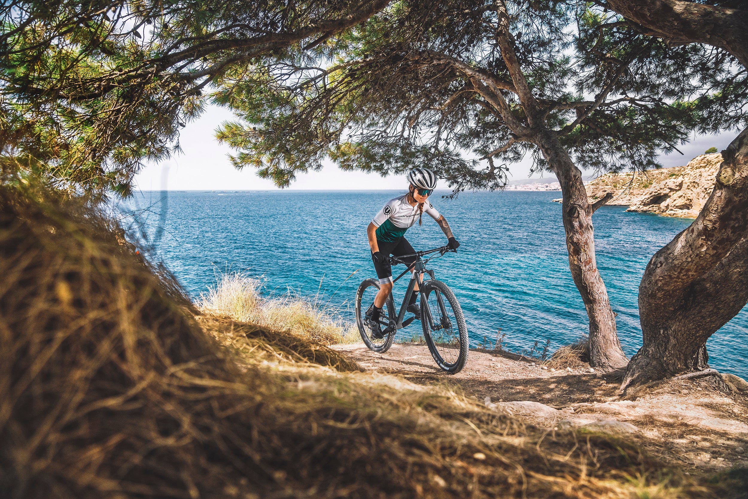 Mountainbiker auf einem Mondraker Podium Rad, fährt auf einem Trail am Meer entlang, umgeben von Bäumen.
