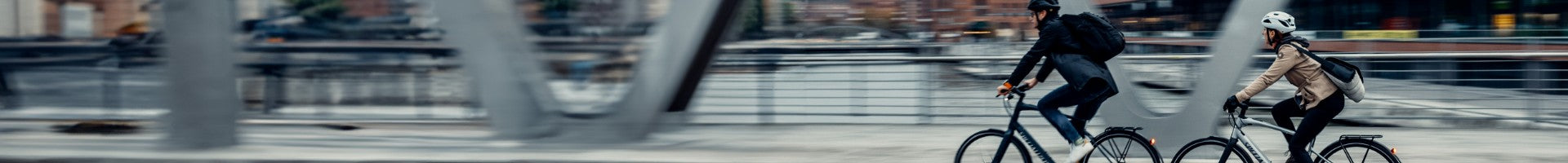 Zwei Personen fahren mit Specialized City- und Urban-Bikes über eine Brücke in der Stadt, beide mit Helm und Rucksack.