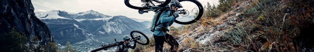Zwei Mountainbiker tragen ihre Fahrräder einen steilen Berghang hinauf; im Hintergrund ist eine alpine Landschaft zu sehen.