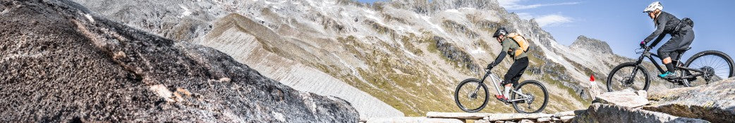 Zwei Mountainbiker fahren auf einem steinigen Pfad in alpiner Landschaft.