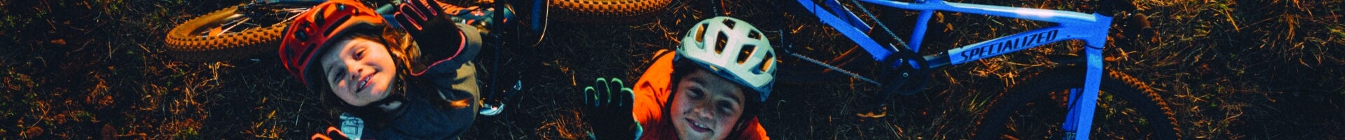 Kinder mit Helmen neben einem blauen Specialized-Fahrrad im Freien, lachen in die Kamera.