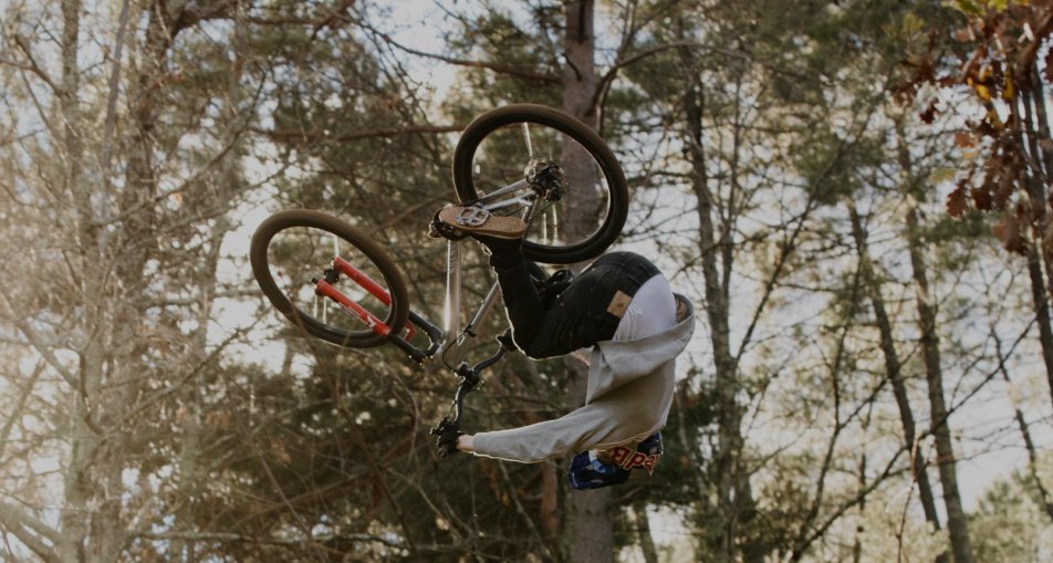 Person führt BMX-Stunt in der Luft im Wald aus, trägt Schutzkleidung und Helm; Fahrrad hat rote Gabel.