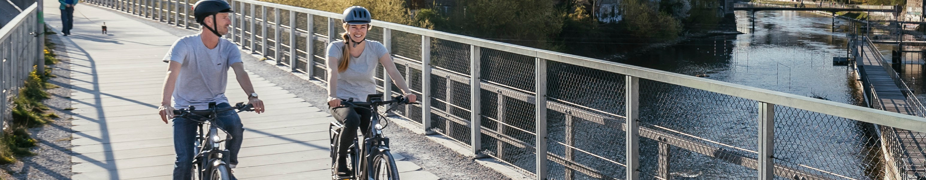 Zwei Personen fahren E-Bikes auf einer Brücke an einem Fluss entlang. Beide tragen Helme.