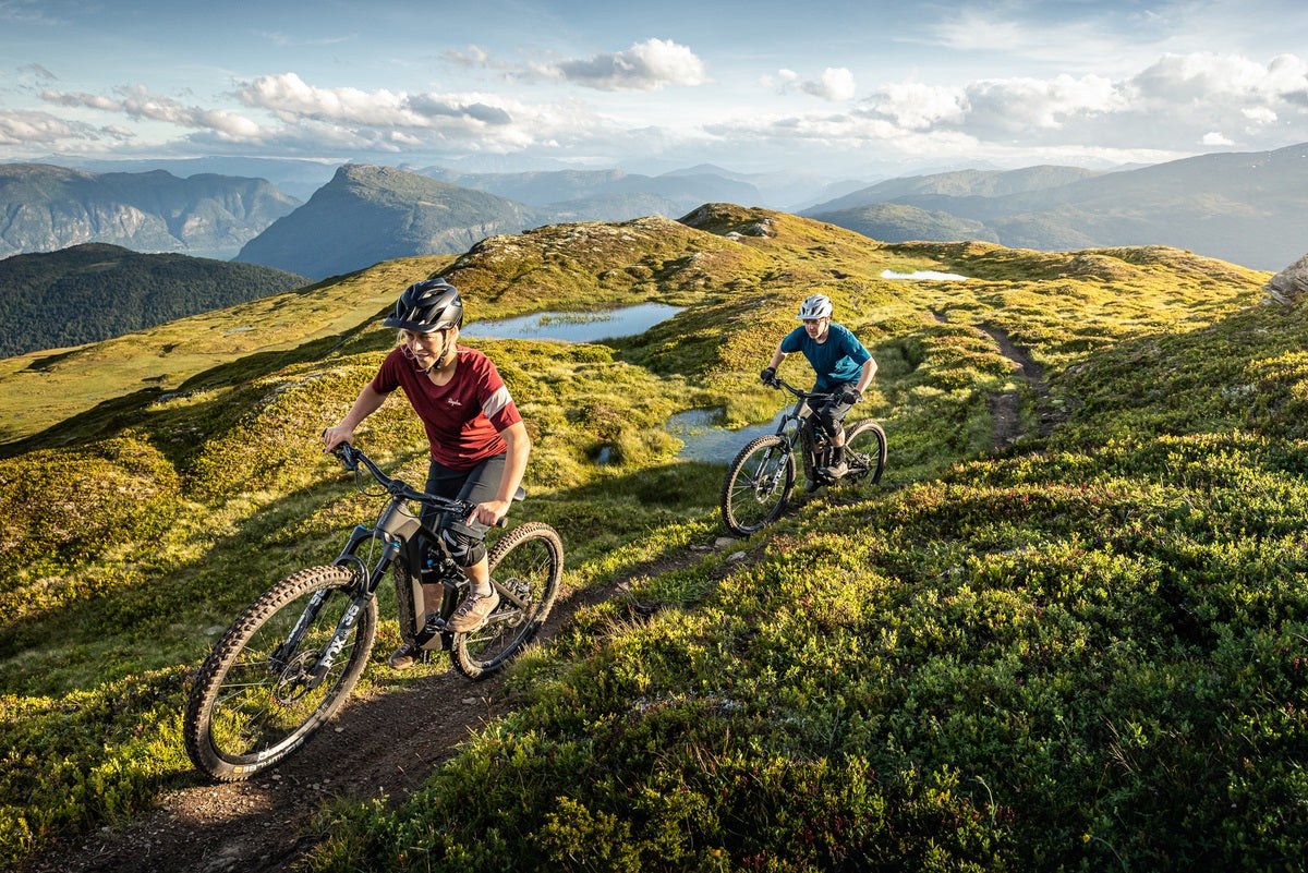 Zwei Personen fahren mit Focus E-Mountainbikes auf einem schmalen Pfad in einer bergigen Landschaft bei Sonnenschein.