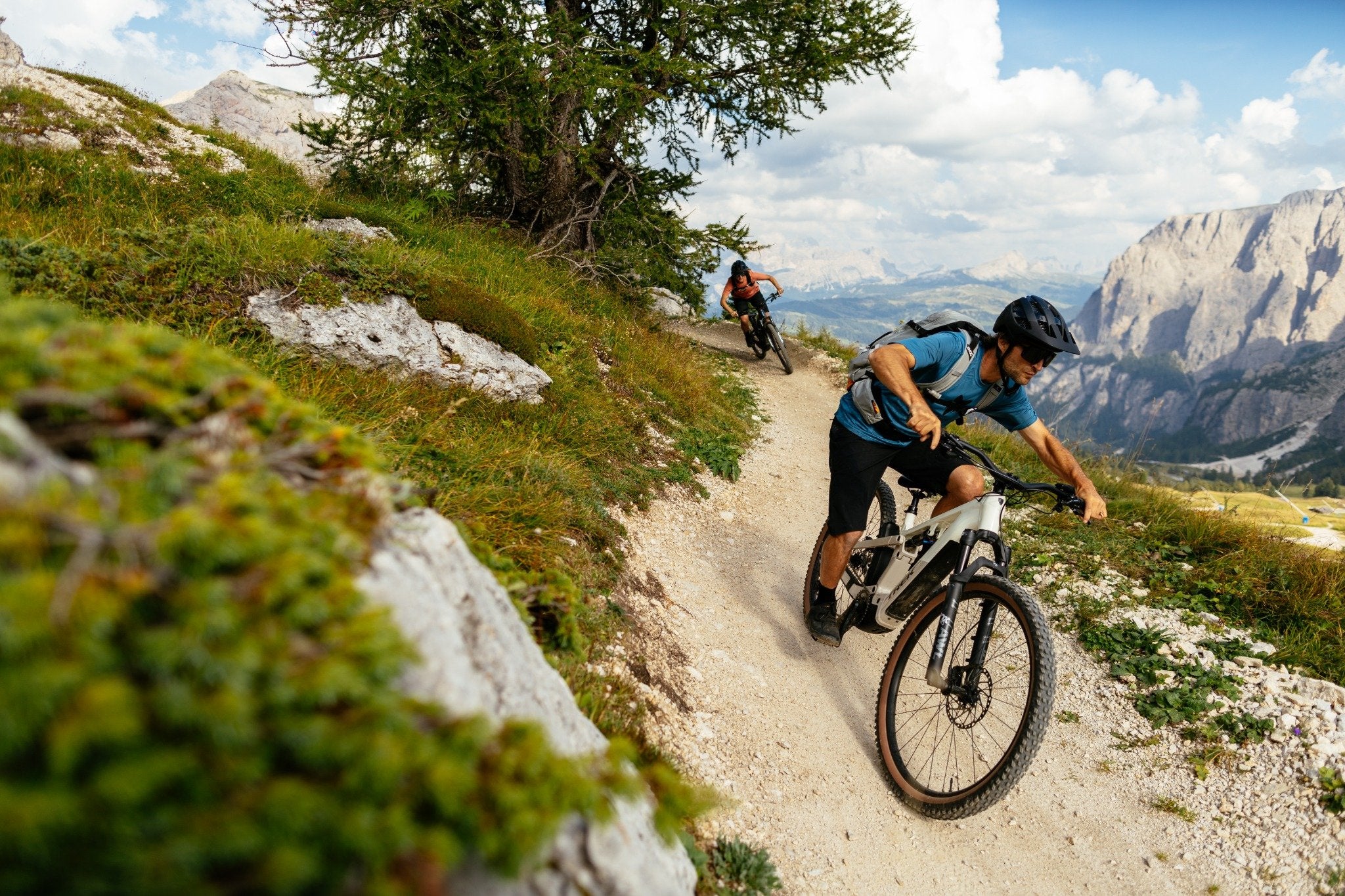Zwei Mountainbiker fahren mit Trek Powerfly FS E-Bikes auf einem schmalen Bergpfad durch eine alpine Landschaft.