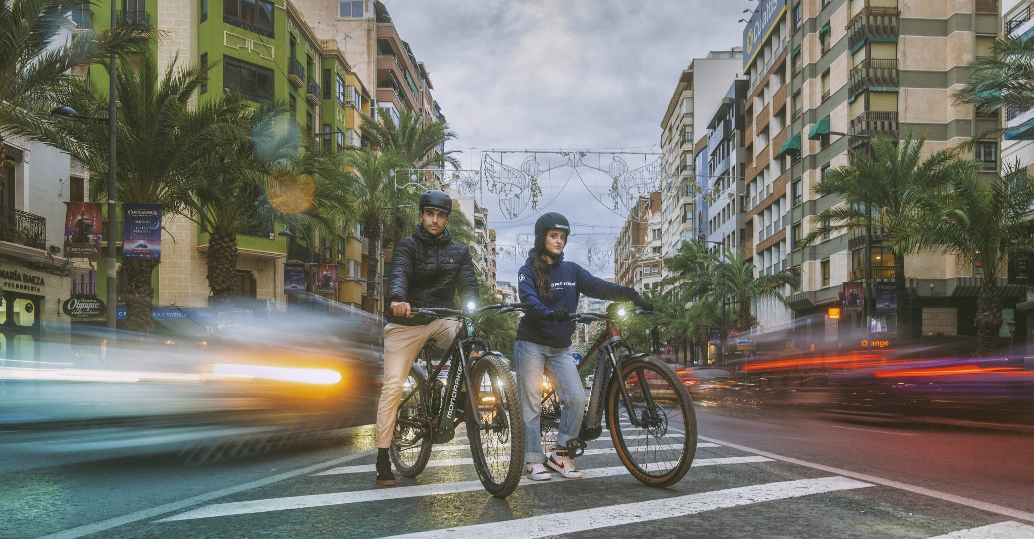 Zwei Personen auf Mondraker Fahrrädern auf einer Straße in der Stadt, umgeben von Verkehr und Beleuchtung.