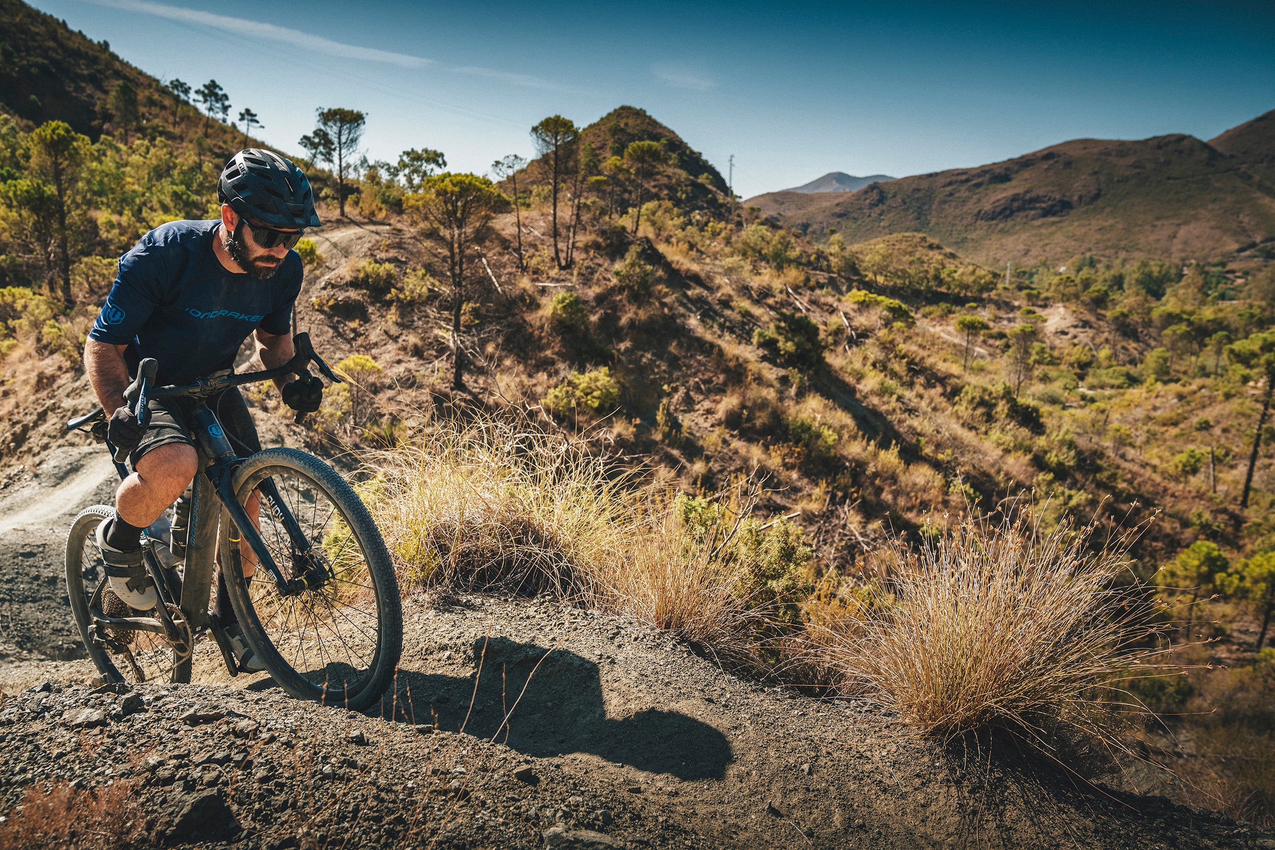 Mann auf Mondraker Dusty Gravelbike im hügeligen Gelände bei sonnigem Wetter, trägt Schutzhelm und Sportbekleidung.