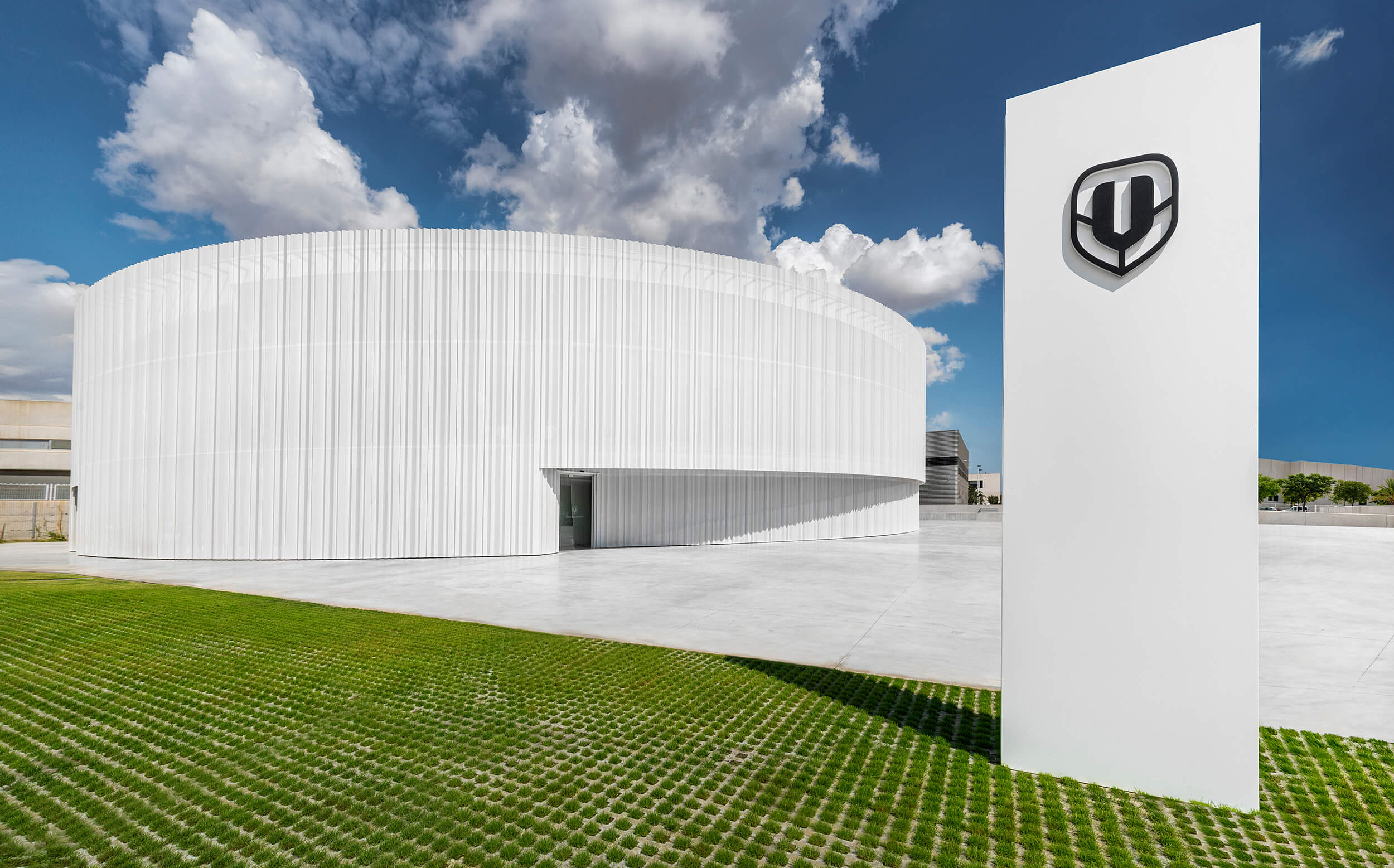 Moderner, weißer Gebäudekomplex mit Logo auf Säule und grünem Rasen davor, unter blauem Himmel.