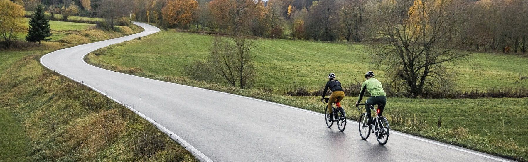 Zwei Personen auf Rennrädern fahren eine kurvenreiche Landstraße entlang, umgeben von herbstlicher Landschaft.