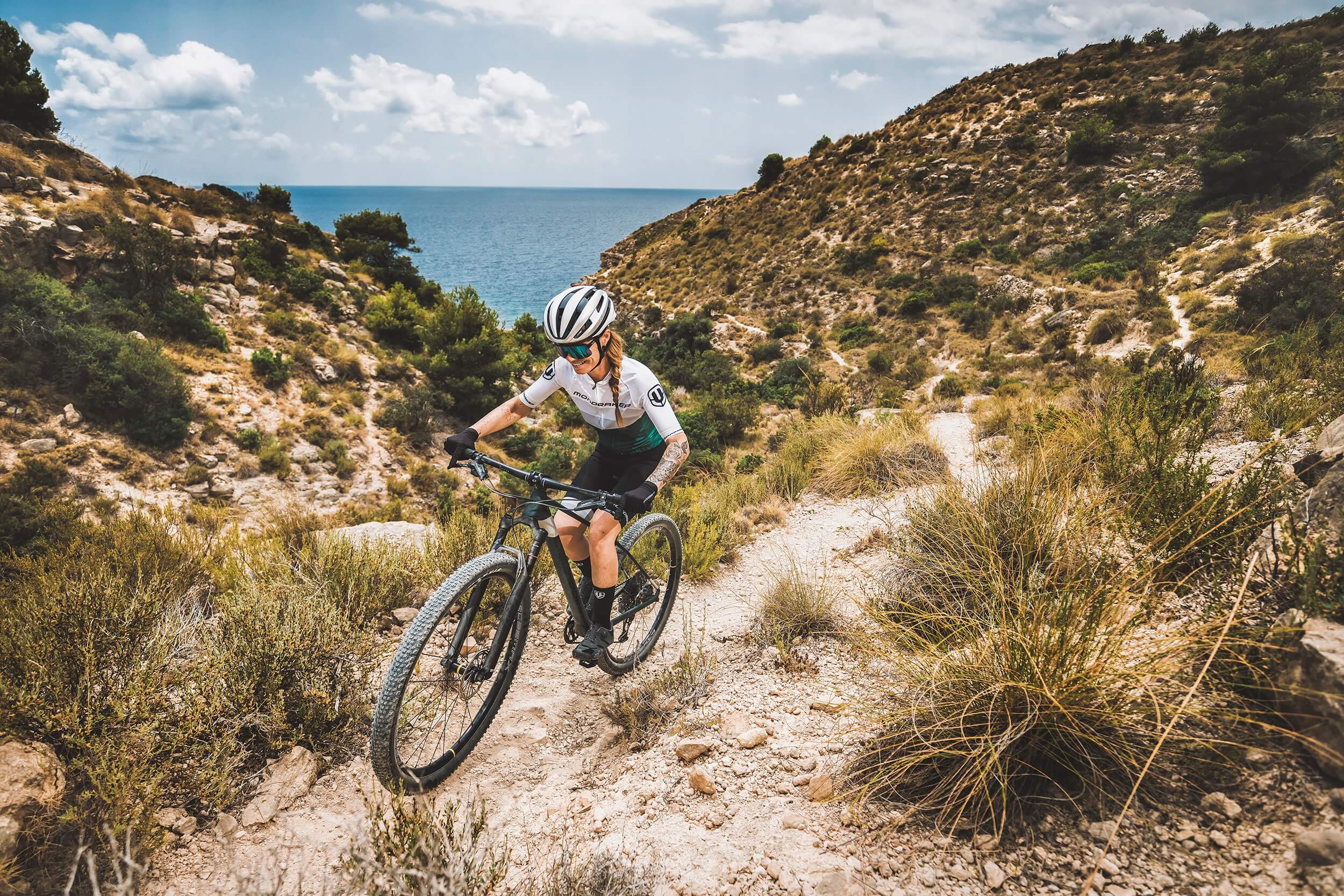 Frau fährt Mondraker-Hardtail-Mountainbike auf steinigem Pfad nahe Küste, ideal für Outdoor- und Bike-Enthusiasten.